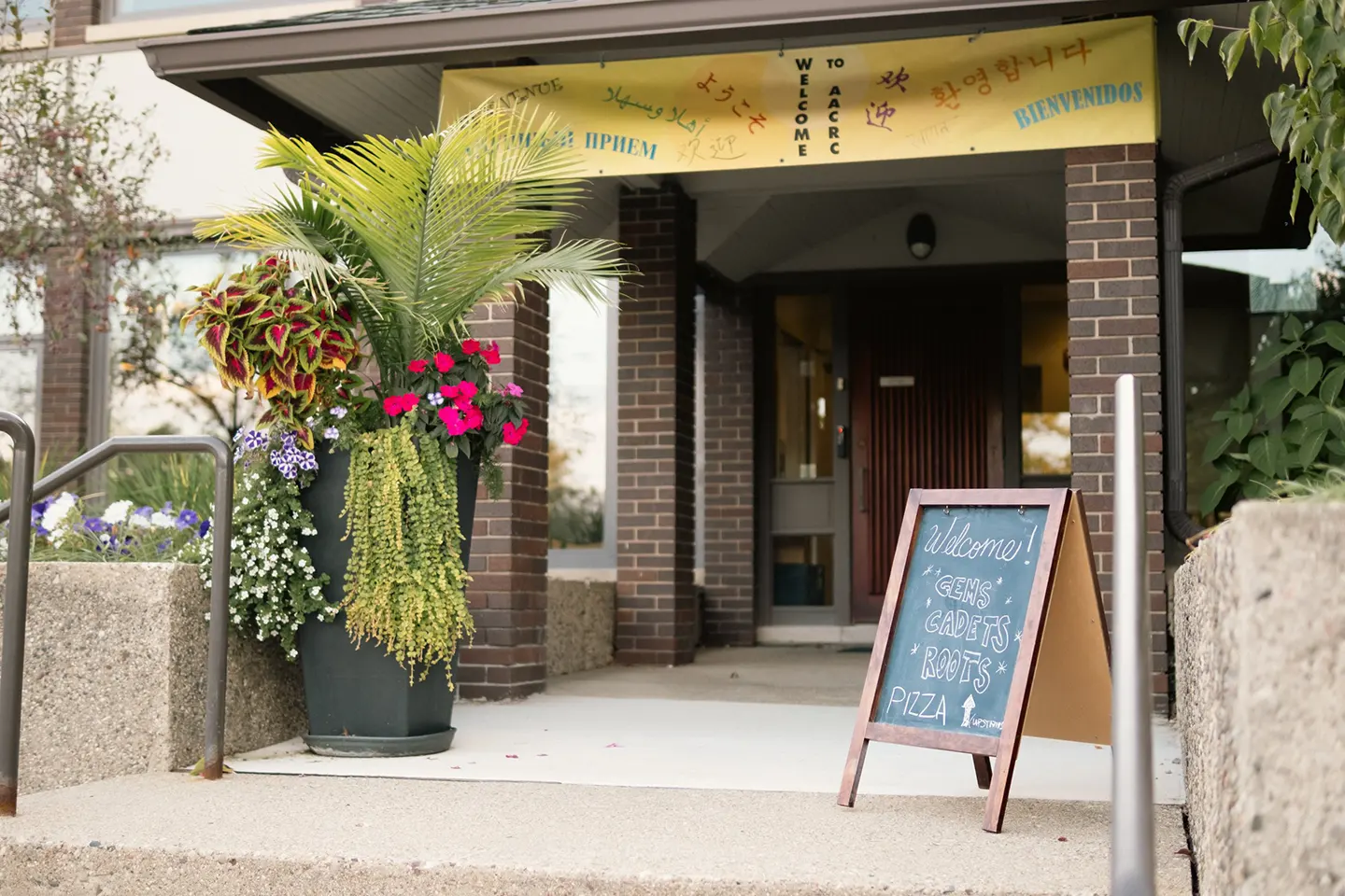 The church entrance, with a multilingual welcome banner above the door, planters of flowers, and a chalkboard sign listing programs