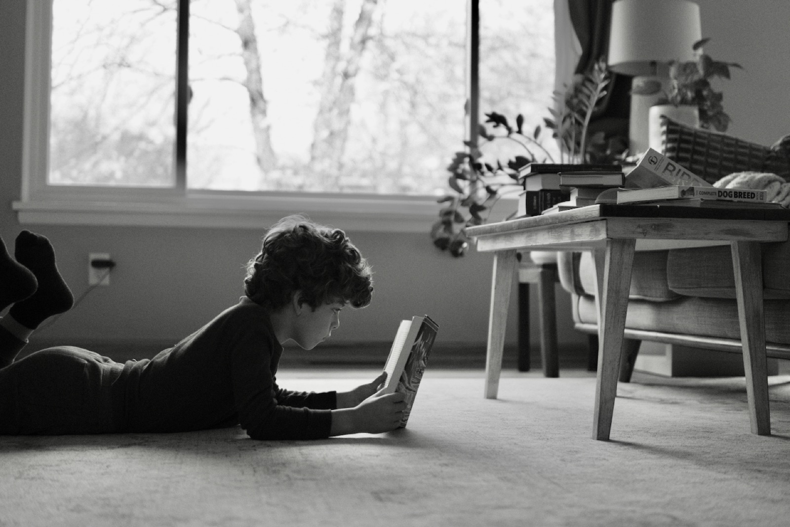 A boy lies on the floor reading a book by window light, black and white