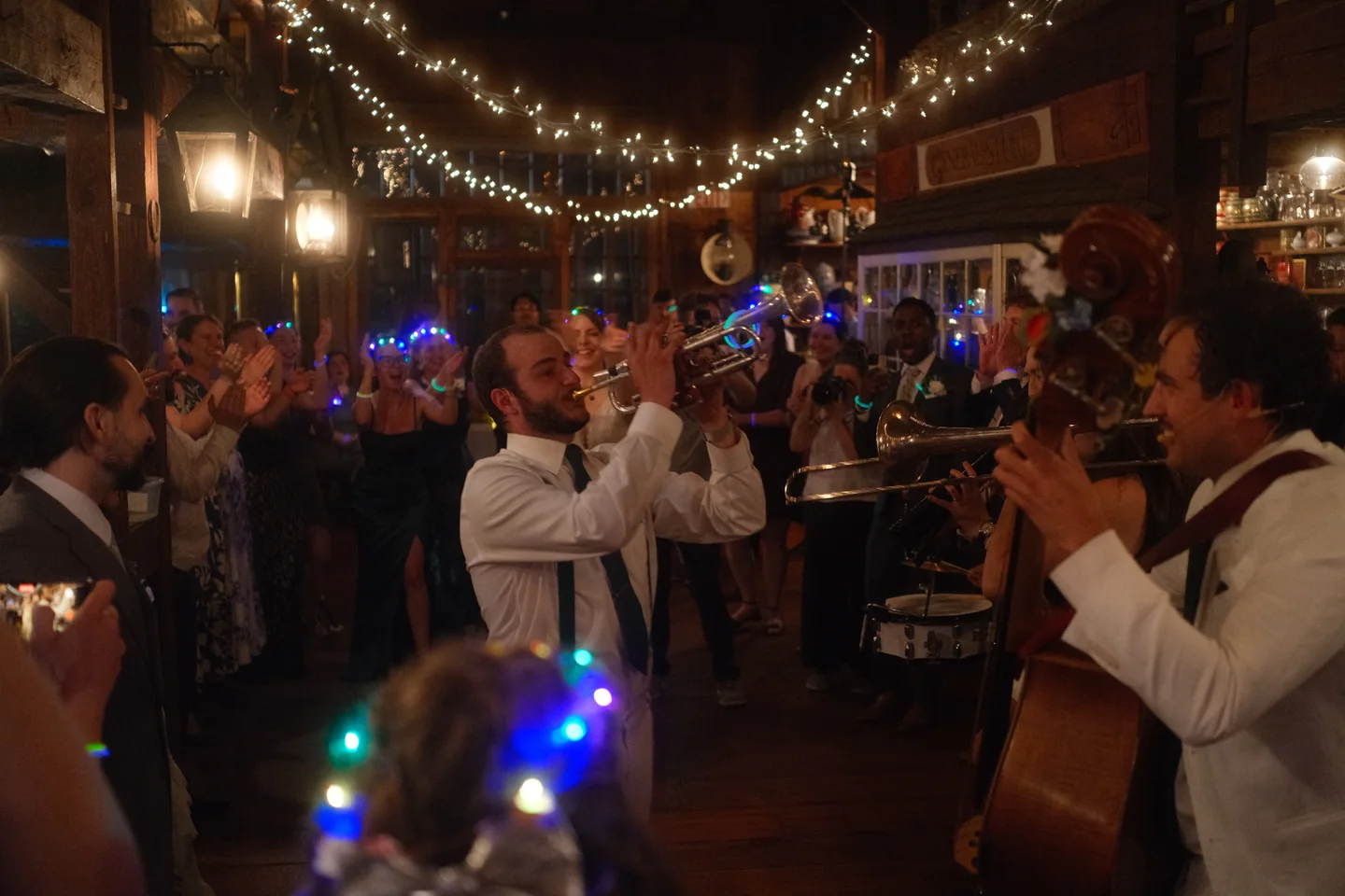 A brass band plays under string lights, trumpet and trombone raised, dancers crowding in at the edges of the frame